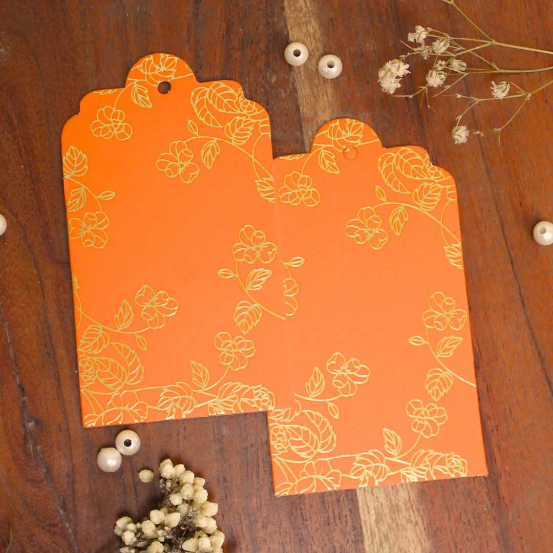 Two orange gift tags with a floral foiled pattern placed on a wooden surface surrounded by decorative elements.